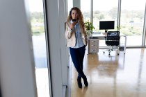 Joven mujer de negocios hablando en el teléfono inteligente mientras se apoya en la ventana en la oficina creativa. Negocios, trabajo de oficina y tecnología inalámbrica. - foto de stock