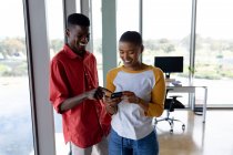 Smiling african american businesswoman showing smartphone to male colleague in creative office. creative business, wireless technology and office workplace. — Stock Photo