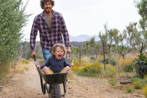 Jovem empurrando alegre e animado filho sentado no carrinho de mão na passarela na fazenda. família, propriedade e prazer. — Fotografia de Stock