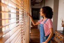 Thoughtful biracial woman in bib overalls looking through window blinds at home. domestic lifestyle and spending time at home. — Stock Photo