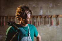 Retrato de herrero barbudo confiado sosteniendo el martillo mientras está parado en la industria. forja, metalurgia e industria manufacturera. - foto de stock