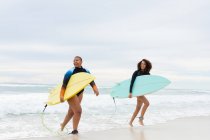 Sonrientes amigas multirraciales con tablas de surf corriendo en la orilla del mar contra el cielo durante el fin de semana. amistad, surf y tiempo libre. - foto de stock