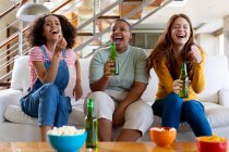 Multiracial female friends with beer and popcorn laughing while watching tv at home. friendship, socialising and leisure time at home. — Stock Photo
