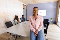 Портрет улыбающейся афроамериканской бизнесвумен в повседневной жизни, сидящей на столе для совещаний в офисе. креативный бизнес и современный офис. — стоковое фото