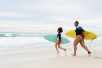 Longitud completa de amigas multirraciales despreocupadas con tablas de surf corriendo en la playa durante el fin de semana. amistad, surf y tiempo libre. - foto de stock