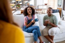 Joyeuses amies multiraciales prenant un café assis sur le canapé à la maison. amitié, socialisation et loisirs à la maison. — Photo de stock