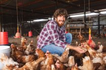 Portrait d'un jeune homme barbu souriant accroupi tout en nourrissant les poules de pastilles en enclos à la ferme. exploitation familiale et avicole, élevage. — Photo de stock