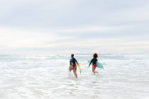 Unbekümmerte multirassische Freundinnen mit Surfbrettern, die am Wochenende im Meer gegen den Himmel laufen. Freundschaft, Surfen und Freizeit. — Stockfoto