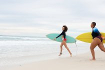Despreocupadas amigas multirraciales con tablas de surf corriendo en la playa durante el fin de semana. amistad, surf y tiempo libre. - foto de stock
