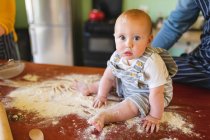 Портрет милого ребенка, играющего, сидя на муке за столом с родителями на кухне. невиновность и семья. — стоковое фото