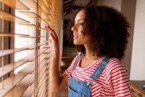 Thoughtful biracial young woman in bib overalls looking through window blinds at home. domestic lifestyle and spending time at home. — Stock Photo