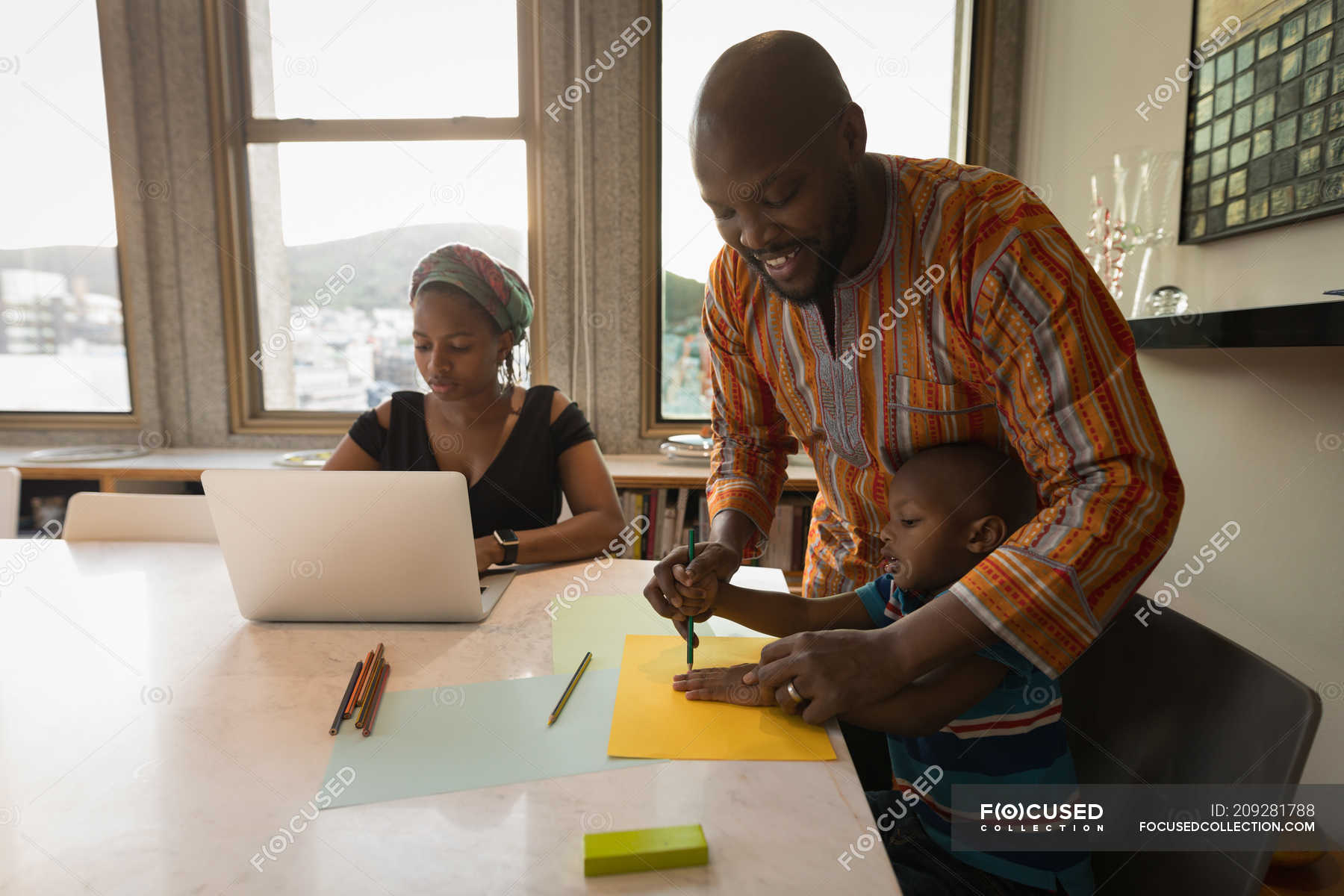 Father Assisting Son Drawing Sketch At Table With Mother Using