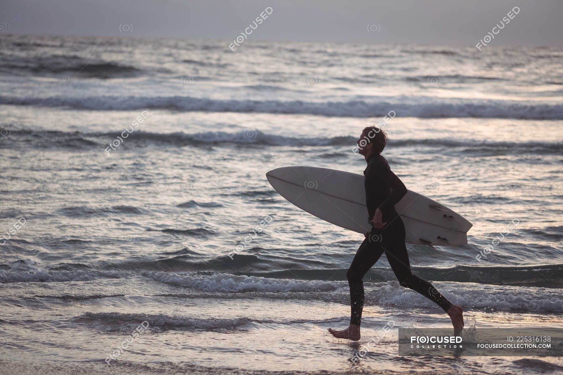 Side view of a man carrying surfboard running on beach at dusk ...