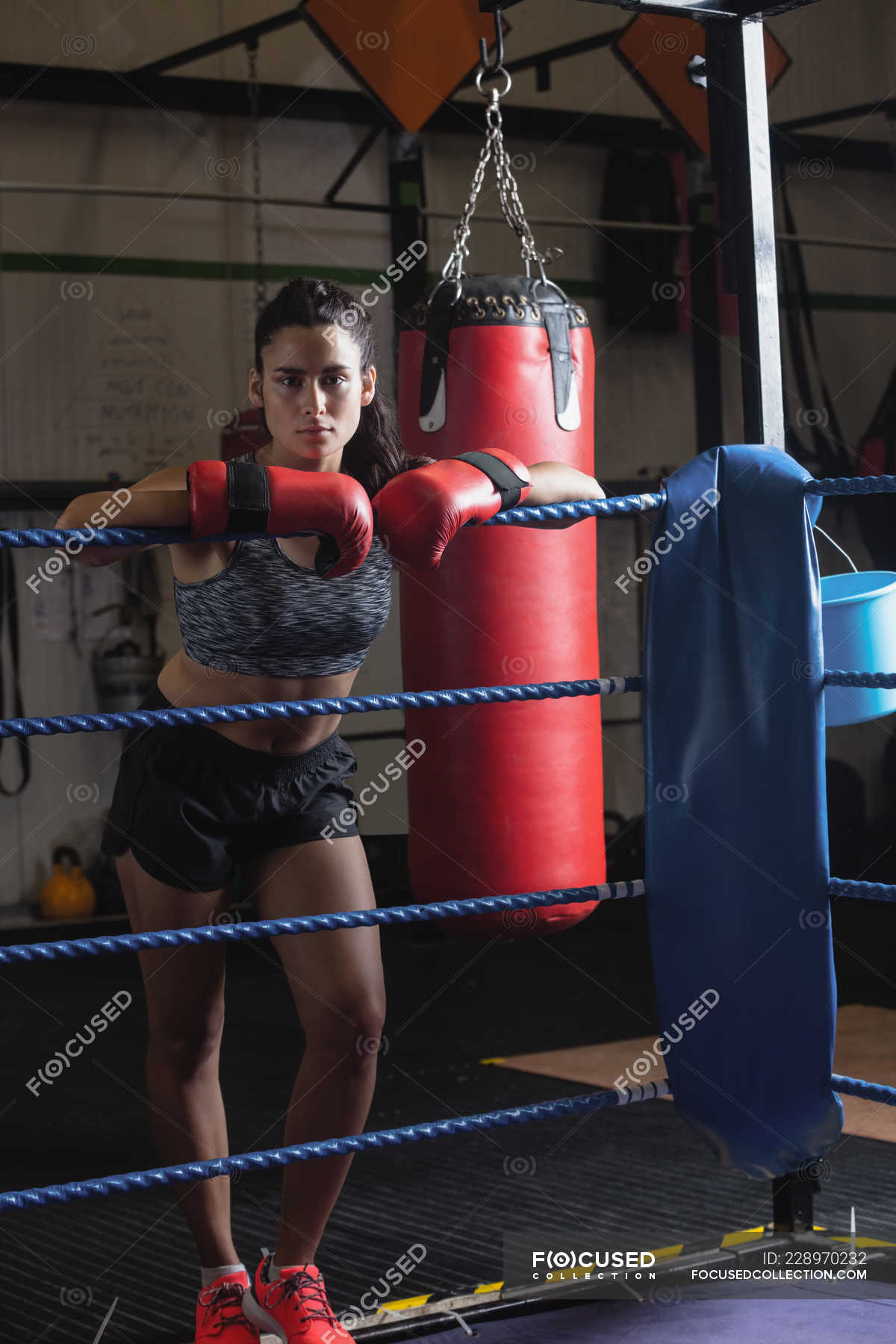 Portrait of female boxer in boxing gloves leaning on boxing ring rope ...