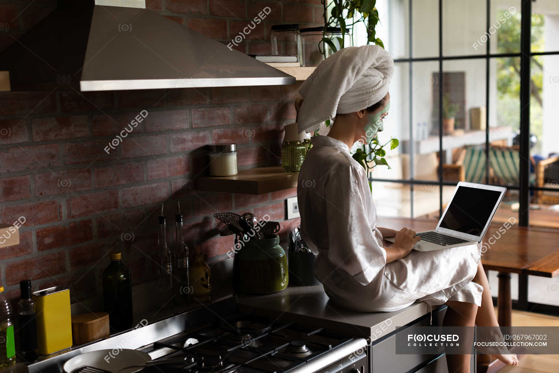 Woman in bathrobe sitting on the kitchen counter and using laptop in ...