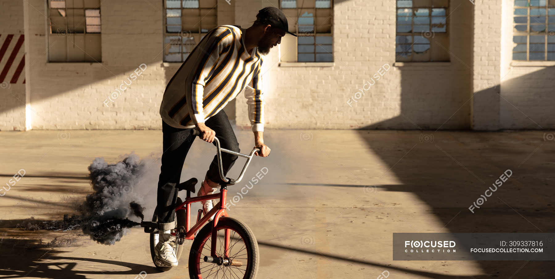 Front view of a young Caucasian man riding a BMX bike with a grey smoke  grenade attached to it, in an abandoned warehouse — unaccompanied, extreme  sport - Stock Photo | #309337816