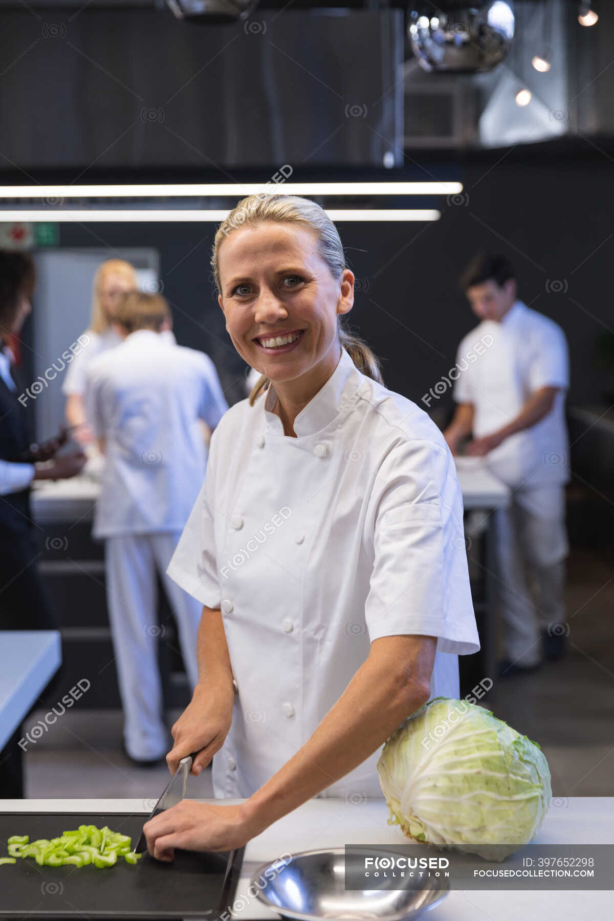 Portrait Of A Caucasian Female Chef Cutting Vegetables Looking At The Camera And Smiling With Other Chefs Cooking In The Background Cookery Class At A Restaurant Kitchen Workshop Happy Stock