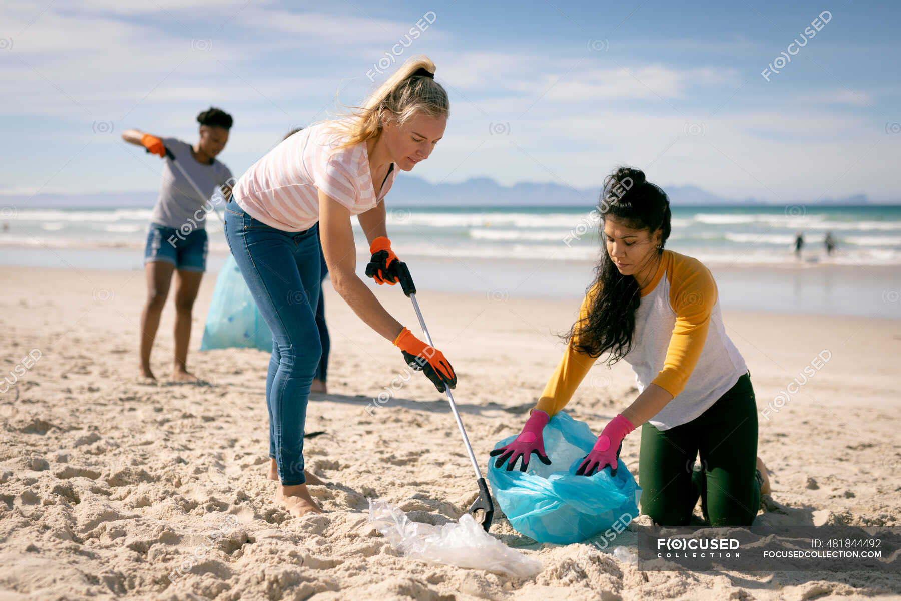 Diverse group of women walking along beach, picking up rubbish. eco ...