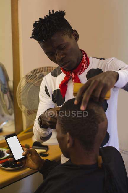 Cliente utilizzando il telefono cellulare mentre il barbiere tagliare i capelli in negozio di barbiere — Foto stock