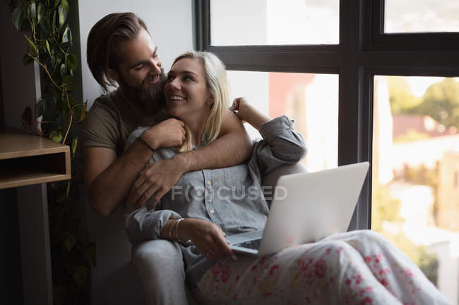 Pareja abrazándose entre sí mientras se utiliza el ordenador portátil en casa - foto de stock