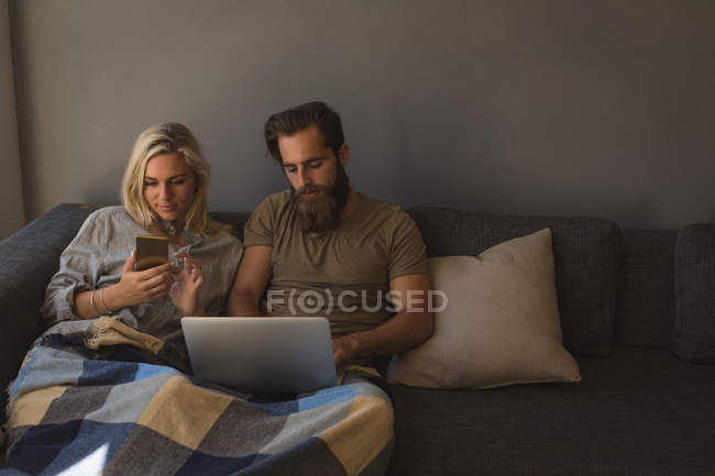 Couple utilisant ordinateur portable et téléphone portable dans le salon à la maison — Photo de stock