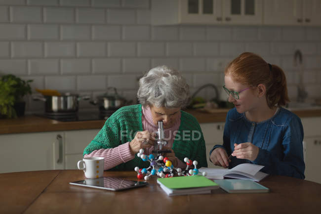 Grand-mère avec sa petite-fille regardant au microscope dans la cuisine à la maison — Photo de stock