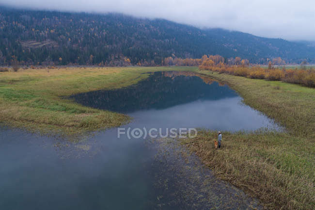 Tourist steht mit Hund in der Nähe eines Sees auf dem Land — Stockfoto