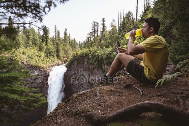 Man sitting near waterfall and drinking water on a sunny day — Stock Photo