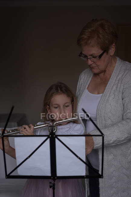 Chica tocando flauta con su abuela en la sala de estar en casa - foto de stock
