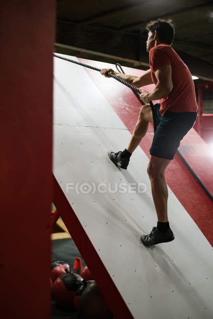 Muskulöser Mann klettert in der Turnhalle an einer Schrägwand mit Seil — Stockfoto