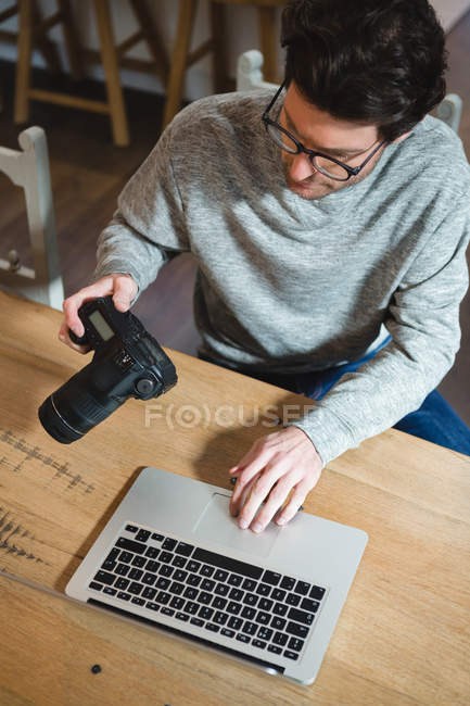 Hombre usando el ordenador portátil mientras sostiene la cámara digital en casa - foto de stock