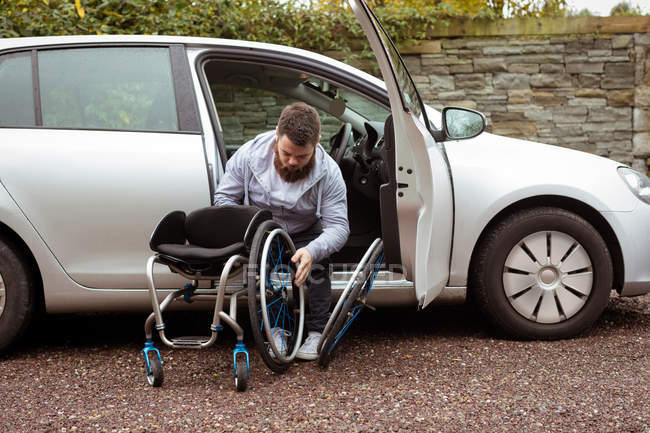 Инвалид молодой человек держит инвалидное кресло, выходя из машины — стоковое фото