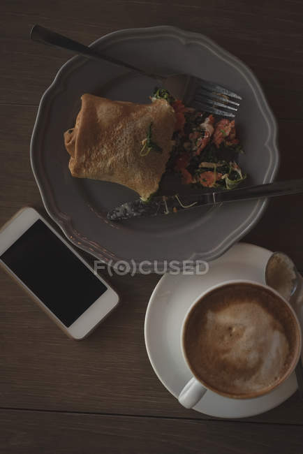 Над чашкою капучино з обгорткою їжі та мобільним телефоном на столі — стокове фото