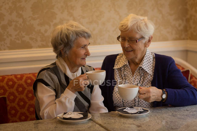 Старші друзі взаємодіють один з одним, маючи каву вдома — стокове фото