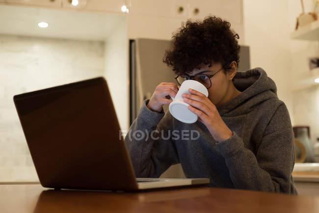 Bella donna che utilizza il computer portatile mentre prende il caffè in cucina — Foto stock
