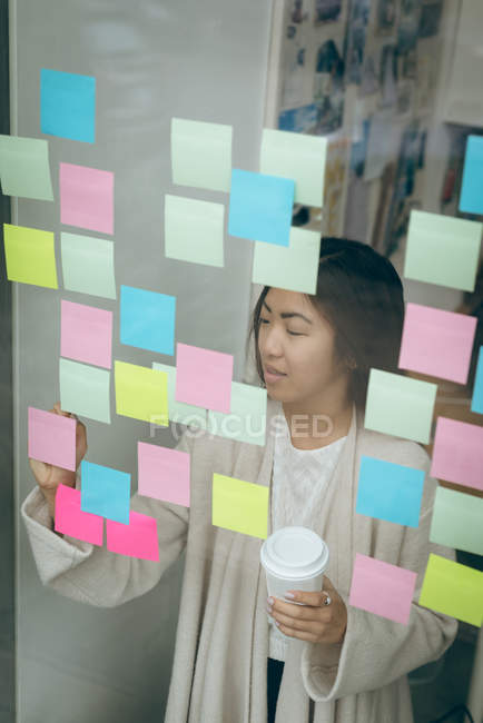 Female executive sticking adhesive notes on glass wall in office — Stock Photo