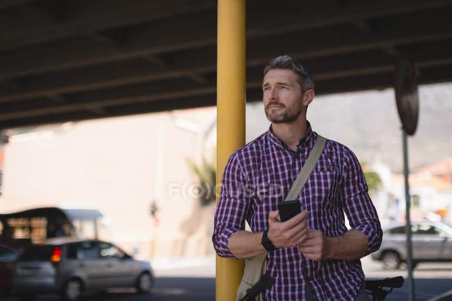 Nachdenklicher Mann hält Handy in der Hand, während er sich an Stange lehnt — Stockfoto