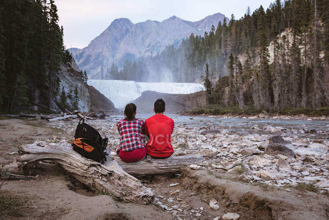Rear view of couple sitting on log near waterfall — Stock Photo