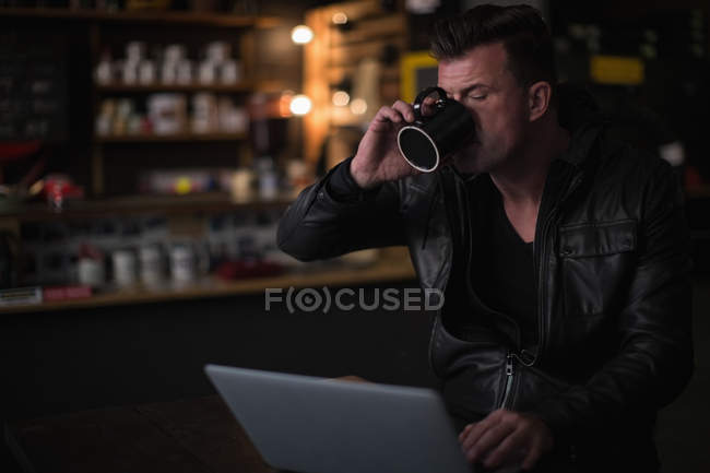 Mecánico usando el ordenador portátil mientras toma café en el garaje - foto de stock