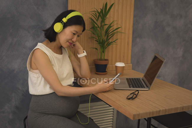 Grávida empresária ouvir música no telefone celular na mesa no escritório — Fotografia de Stock