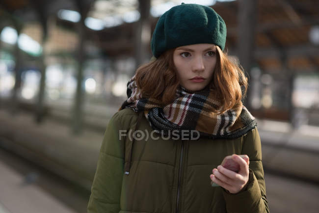 Thoughtful woman in winter clothing holding mobile phone in platform — Stock Photo