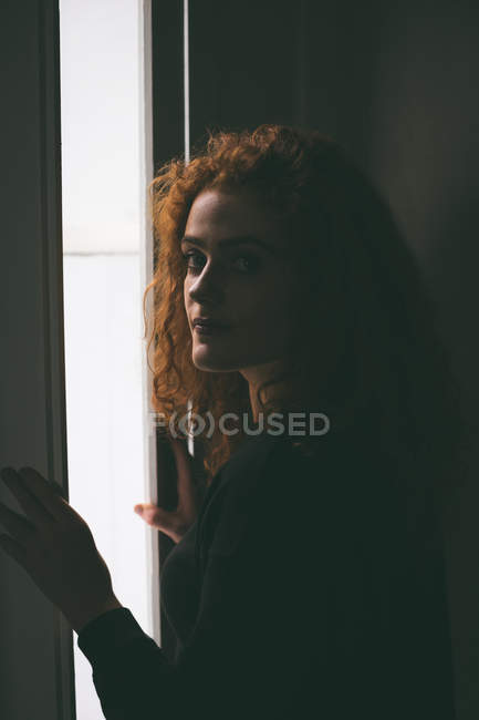 Bella donna guardando la fotocamera mentre in piedi vicino alla finestra — Foto stock