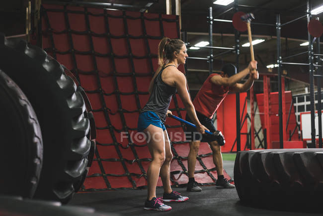 Muskulöses Paar schlägt mit Vorschlaghammer in Turnhalle auf Reifen ein — Stockfoto