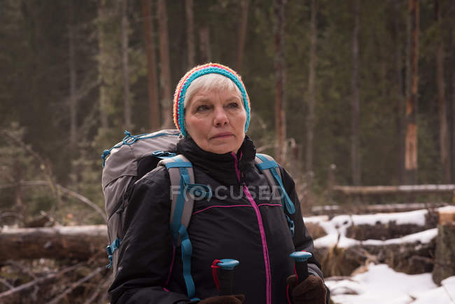 Задумчивая пожилая женщина, стоящая с рюкзаком и походным столбом зимой — стоковое фото
