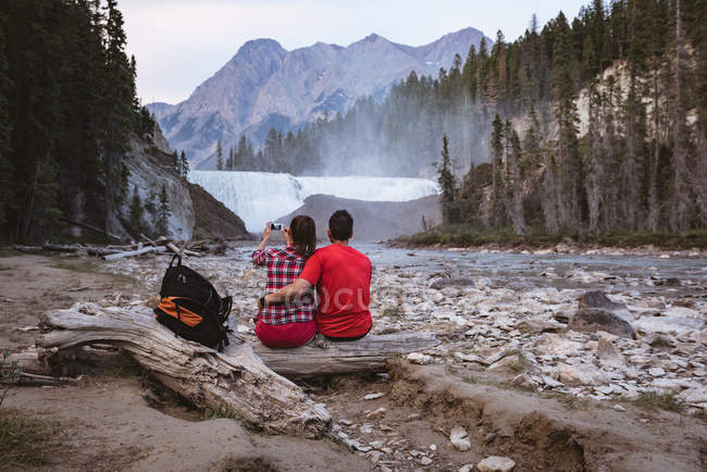 Rear view of couple taking photo of waterfall with mobile phone — Stock Photo