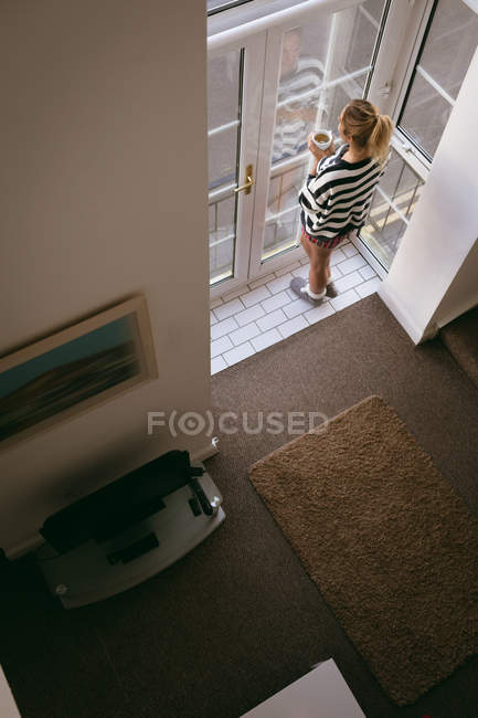 Mulher olhando pela janela enquanto toma café em casa — Fotografia de Stock