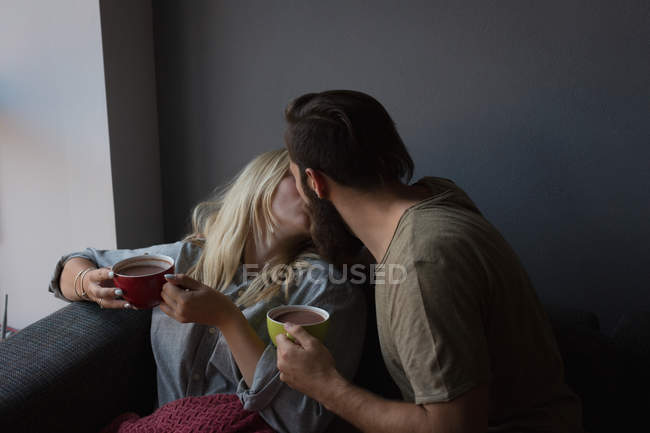 Casal beijando uns aos outros enquanto toma café na sala de estar em casa — Fotografia de Stock