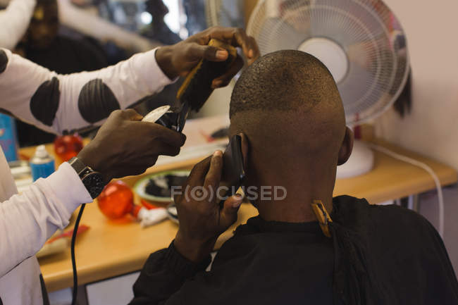 Client parlant sur téléphone portable tout barbier couper ses cheveux dans le salon de coiffure — Photo de stock