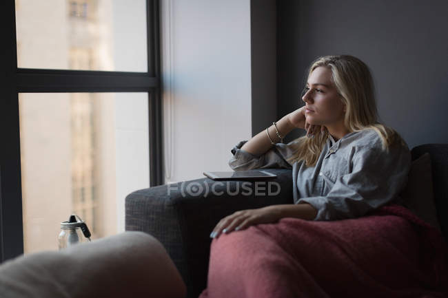 Mujer pensativa sentada con tableta digital en la sala de estar en casa - foto de stock