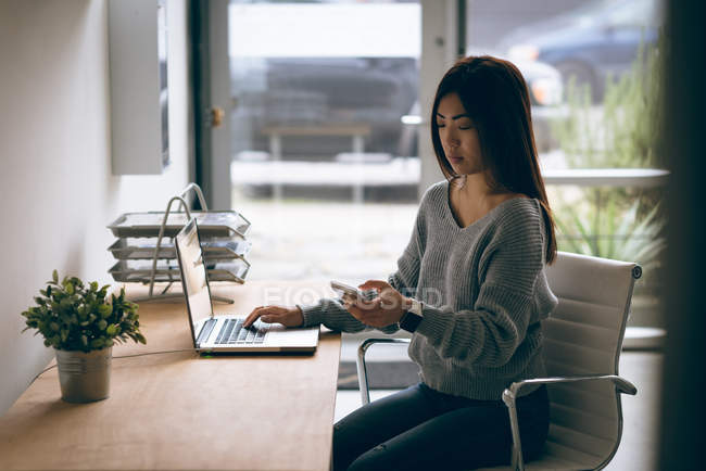 Жіночий керівник використовує мобільний телефон під час роботи на ноутбуці за столом в офісі — стокове фото